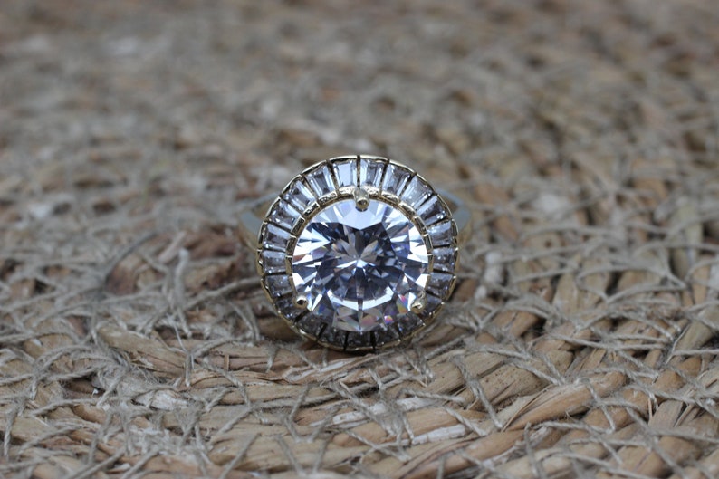 K&ouml;nnte beinhalten: Ein goldener Ring mit einem gro&szlig;en, klaren, runden Edelstein, der von einem Heiligenschein aus kleineren, baguettef&ouml;rmigen Edelsteinen umgeben ist.