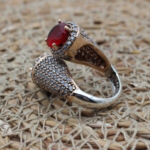 May include: A silver ring featuring a red gemstone and clear stones. The ring is designed with two prongs that wrap around the finger.