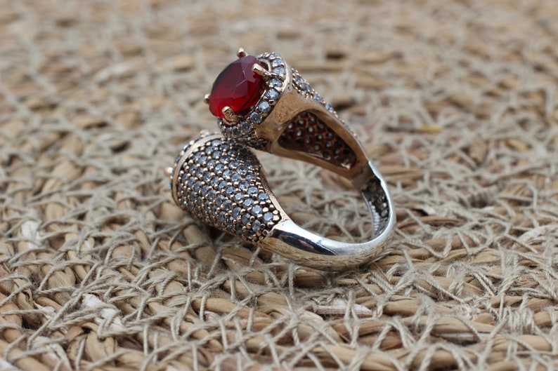 May include: A silver ring featuring a red gemstone and clear stones. The ring is designed with two prongs that wrap around the finger.
