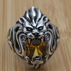 May include: A silver-colored ring featuring a detailed lion's head design. The lion's face has an open mouth with visible teeth, and a large, oval-shaped, yellow gemstone is set in the center. The ring is on a wooden surface.