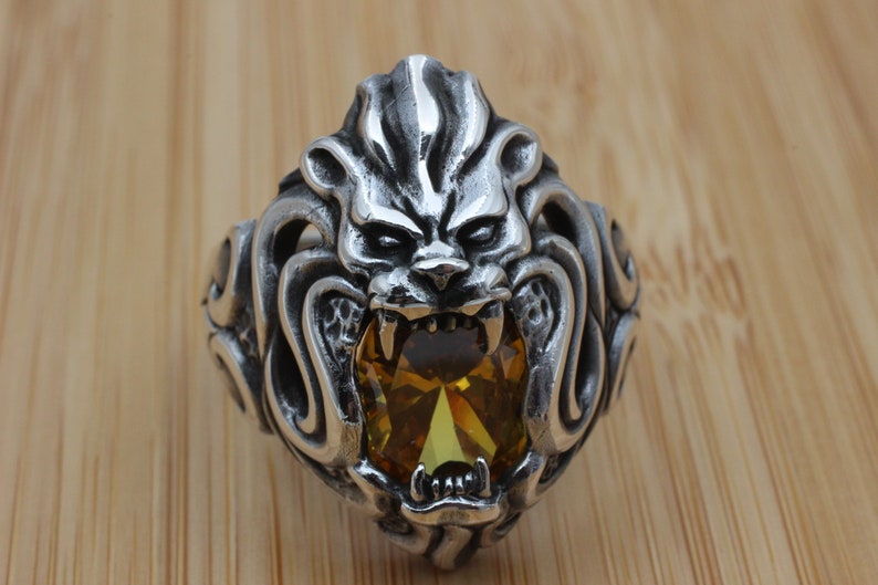 May include: A silver-colored ring featuring a detailed lion's head design. The lion's face has an open mouth with visible teeth, and a large, oval-shaped, yellow gemstone is set in the center. The ring is on a wooden surface.