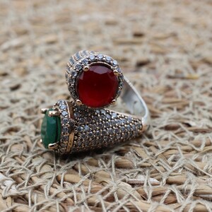 May include: A silver ring featuring two large stones, one red and one green, encircled by smaller clear stones.