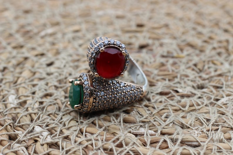 May include: A silver ring featuring two large stones, one red and one green, encircled by smaller clear stones.
