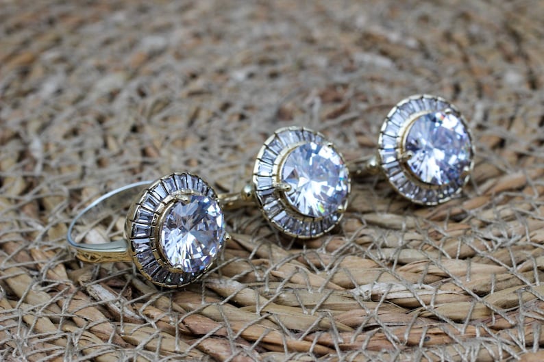 May include: Three gold rings with large clear cubic zirconia stones set in a halo of smaller stones. The rings are on a woven brown background.