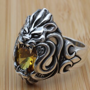 May include: A silver-colored ring featuring a detailed lion's head with an open mouth, revealing sharp teeth. The lion's head holds a large, yellow-green gemstone. The ring has an ornate design on the band.