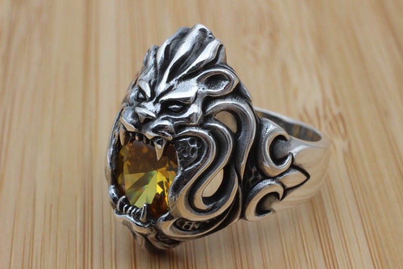 May include: A silver-colored ring featuring a detailed lion's head with an open mouth, revealing sharp teeth. The lion's head holds a large, yellow-green gemstone. The ring has an ornate design on the band.