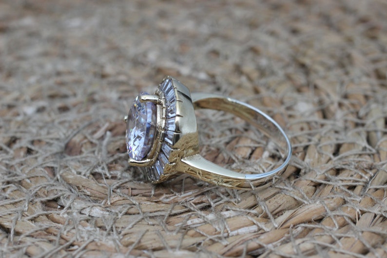 K&ouml;nnte beinhalten: Ein goldener Ring mit einem gro&szlig;en, klaren, ovalen Edelstein, der von einem Heiligenschein aus kleineren, klaren Edelsteinen umgeben ist.