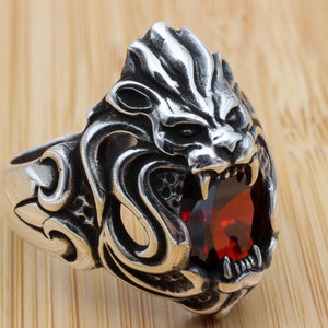 Może przedstawiać: Srebrny pierścionek z detalicznym wzorem głowy lwa. Paszcza lwa jest otwarta, odsłaniając czerwony kamień szlachetny.