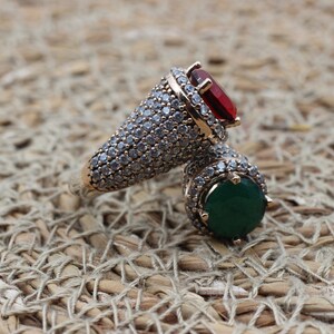 May include: A gold ring with a red and green gemstone setting. The ring is covered in small, clear stones.