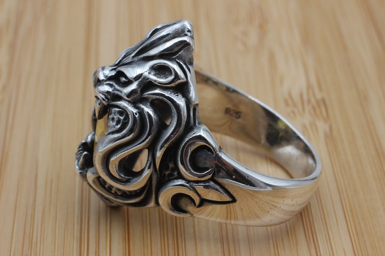 May include: A silver ring featuring a detailed lion's head design. The ring has intricate carvings and a polished finish. The band is marked with "925". This piece is a statement accessory.