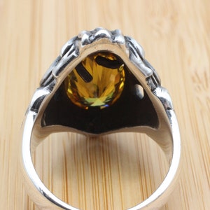 May include: A silver ring with a detailed design around a large, oval, yellow gemstone. The ring's band is smooth and polished, contrasting with the textured setting of the stone. The gemstone has a brilliant cut.