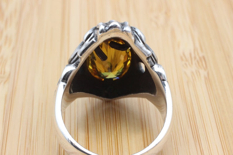 May include: A silver ring with a detailed design around a large, oval, yellow gemstone. The ring's band is smooth and polished, contrasting with the textured setting of the stone. The gemstone has a brilliant cut.