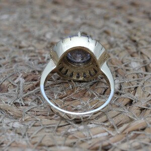 K&ouml;nnte beinhalten: Ein goldener Ring mit einem gro&szlig;en violetten Edelstein, der von kleineren klaren Edelsteinen umgeben ist. Der Ring liegt auf einem gewebten Untergrund.