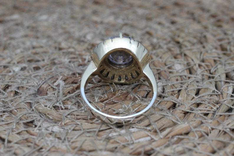 K&ouml;nnte beinhalten: Ein goldener Ring mit einem gro&szlig;en violetten Edelstein, der von kleineren klaren Edelsteinen umgeben ist. Der Ring liegt auf einem gewebten Untergrund.