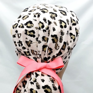May include: A pink and gold leopard print surgical scrub hat with a bright pink ribbon tie. The hat is designed to be worn with a ponytail.