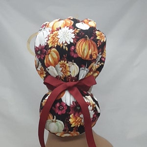 Pode incluir: Uma touca cirúrgica preta com um padrão de outono de abóboras, flores brancas e laranja e detalhes bordô. A touca é amarrada com uma fita bordô. A touca foi projetada para ser usada na cabeça.