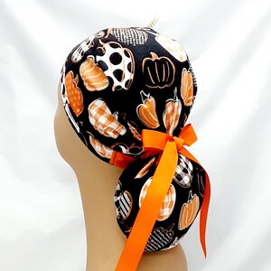 Pode incluir: Um gorro de cirurgia preto com uma estampa de ab&oacute;boras e pimentas brancas e laranja. O gorro tem uma fita laranja longa amarrada em um la&ccedil;o na parte de tr&aacute;s.