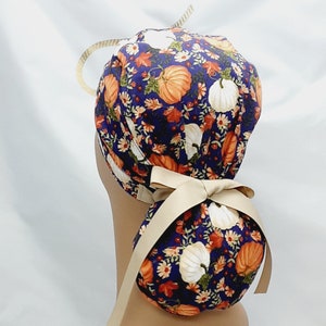 May include: A navy blue scrub cap with a fall-themed pumpkin and floral pattern. The cap features orange and white pumpkins, with a beige ribbon tied in a bow at the back. A gold-colored loop is attached to the top.