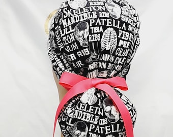 May include: Black and white scrub hat with a pink bow. The hat has a print of anatomical terms and skulls. The text includes "Skeleton", "Mandible", "Ribs", "Patella", "Tibia", "Femur", "Skull", "Pelvis", "Cranium", "Air", and more.