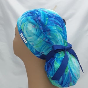 May include: Blue and green surgical scrub hat with a swirling abstract pattern and a blue ribbon tied in a bow at the back. The hat has a white label with the text "Scrub Caps" on it.