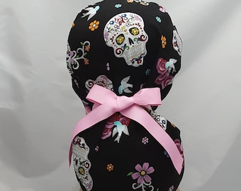 May include: Black scrub hat with a pink bow and a sugar skull pattern. The skull is white with colorful floral details and the background is black with small white flowers and blue birds.