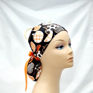 Pode incluir: Um gorro cir&uacute;rgico preto e laranja estampado com um furo para rabo de cavalo e um la&ccedil;o de fita laranja correspondente. O gorro tem um design de ab&oacute;bora.
