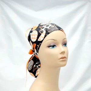 Pode incluir: Um gorro cir&uacute;rgico preto, branco e laranja estampado com um la&ccedil;o preto. O gorro tem um desenho de ab&oacute;boras e outros elementos tem&aacute;ticos de Halloween.