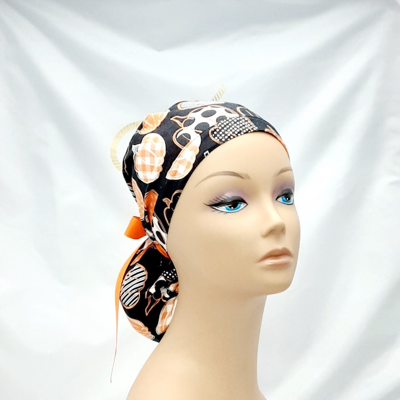 Pode incluir: Um gorro cir&uacute;rgico preto, branco e laranja estampado com um la&ccedil;o preto. O gorro tem um desenho de ab&oacute;boras e outros elementos tem&aacute;ticos de Halloween.