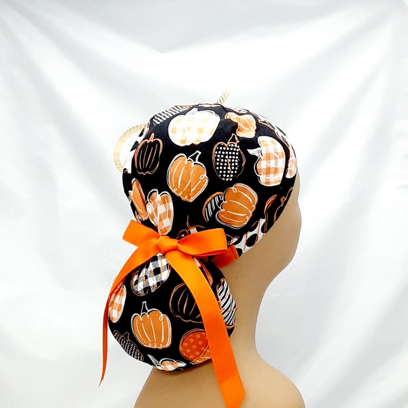 Pode incluir: Um gorro cir&uacute;rgico preto e laranja com um desenho de ab&oacute;bora. O gorro tem uma fita longa laranja amarrada na parte de tr&aacute;s.