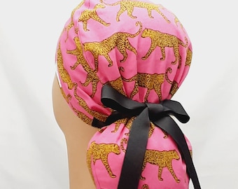 Könnte beinhalten: Rosa chirurgischer Scrubb-Hut mit Leopardenmuster in Gold. Der Hut hat eine schwarze Schleife.