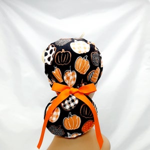 Pode incluir: Um gorro de cirurgia preto e laranja com estampa de ab&oacute;bora e uma fita laranja amarrada em um la&ccedil;o na parte de tr&aacute;s.