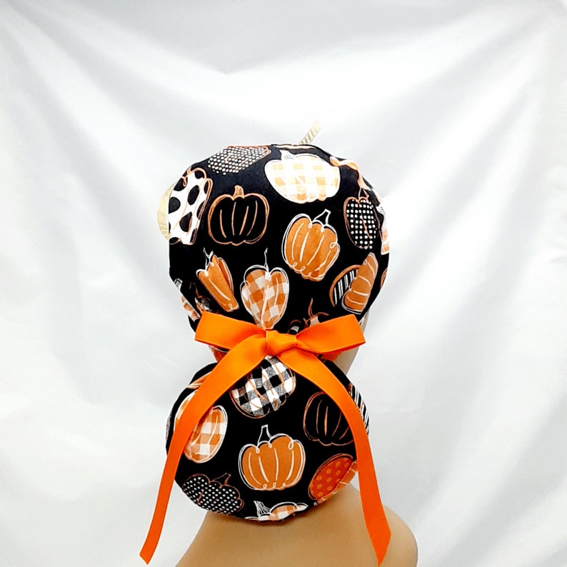 Pode incluir: Um gorro de cirurgia preto e laranja com estampa de ab&oacute;bora e uma fita laranja amarrada em um la&ccedil;o na parte de tr&aacute;s.