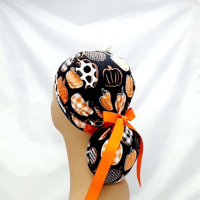Pode incluir: Um gorro de cirurgia preto com uma estampa de ab&oacute;boras e pimentas brancas e laranja. O gorro tem uma fita laranja longa amarrada em um la&ccedil;o na parte de tr&aacute;s.