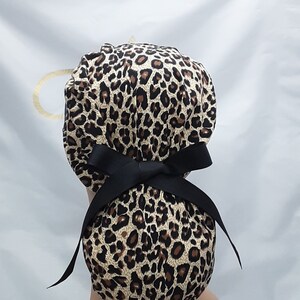 May include: A leopard print surgical scrub hat with a black ribbon tie. The hat is tied in a bow at the back.