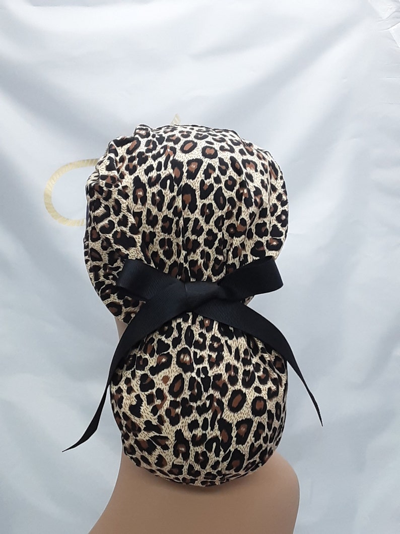 May include: A leopard print surgical scrub hat with a black ribbon tie. The hat is tied in a bow at the back.