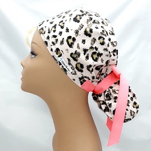 May include: A pink and brown leopard print surgical scrub hat with a pink tie. The hat has the text "Stitch & Hustle" printed on the side.