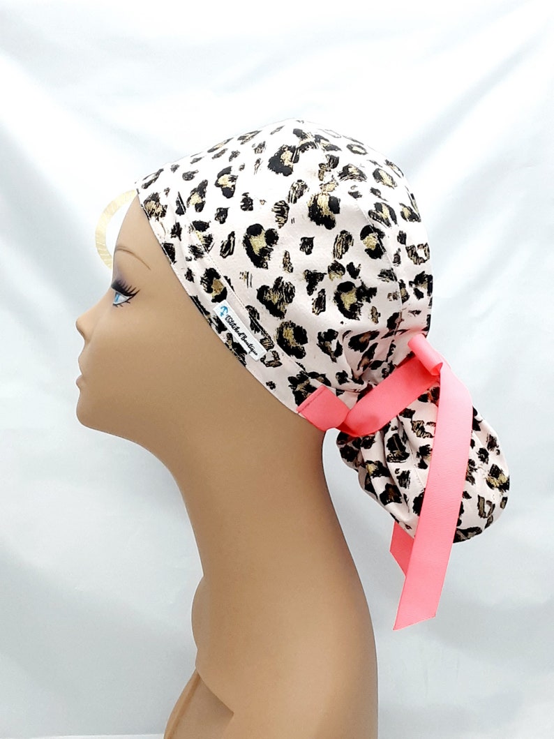 May include: A pink and brown leopard print surgical scrub hat with a pink tie. The hat has the text "Stitch & Hustle" printed on the side.