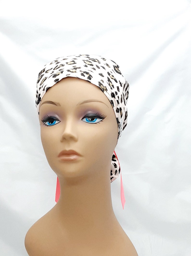 May include: A white and brown leopard print surgical scrub hat with pink ties. The hat is made of a soft, comfortable fabric and is designed to be worn by medical professionals.