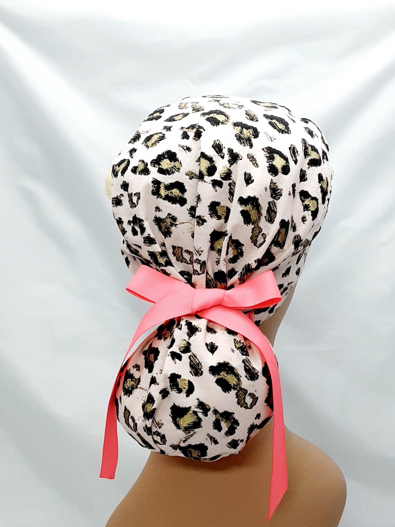 May include: A pink and gold leopard print surgical scrub hat with a bright pink ribbon tie. The hat is designed to be worn with a ponytail.