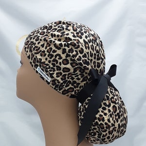May include: A leopard print surgical scrub hat with a black ribbon tie. The hat has a ponytail opening and the text "The Scrub Boutique" is visible on the side.