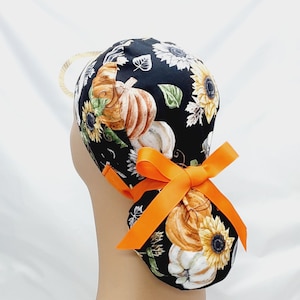 Pode incluir: Uma touca cirúrgica preta com um padrão outonal de abóboras, girassóis e folhas em tons de laranja, branco e verde. A touca é amarrada com uma fita laranja brilhante.