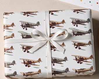 May include: A rectangular gift box wrapped in white paper featuring a repeating pattern of vintage biplane illustrations in shades of brown and gray. The package is tied with a thin, cream-colored satin ribbon tied in a centered bow.