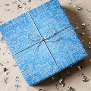May include: A square gift box wrapped in bright blue paper featuring a white circuit board pattern. The box is tied with a thin, silver metallic string arranged in a decorative bow centered on the top surface of the package.