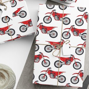 Op de afbeelding: Wit cadeaupapier met een herhaald patroon van rode motorfietsen. De ingepakte cadeaus zijn vastgebonden met touw en versierd met witte labels. Een bol touw en een schaar zijn ook zichtbaar.