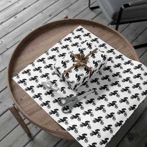 Op de afbeelding: Een ingepakte geschenkdoos met een bruine strik ligt op wit inpakpapier. Het papier en de doos zijn bedekt met een herhalend zwart motorfiets-silhouetpatroon. Schaar en een zilveren lepel liggen ook op het papier.