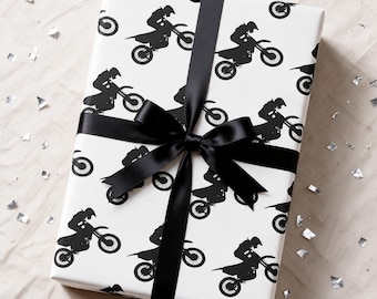 May include: A rectangular gift box wrapped in white paper featuring a repeating black silhouette pattern of riders on motocross bikes. The package is tied with a smooth black satin ribbon that is tied into a centered bow on the top surface.