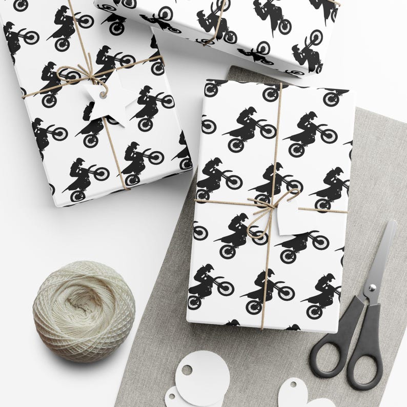 Op de afbeelding: Cadeaudozen verpakt in wit papier met een zwart motorpatroon. De dozen zijn vastgebonden met touw en hebben blanco labels. Een bol touw, een schaar en blanco labels zijn ook zichtbaar.
