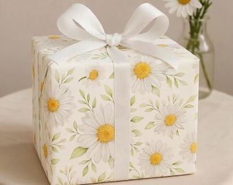 May include: A square gift box wrapped in cream-colored paper featuring a repeating pattern of white daisy flowers with yellow centers and light green leaves. The box is tied with a wide, smooth white satin ribbon tied in a large bow on top.