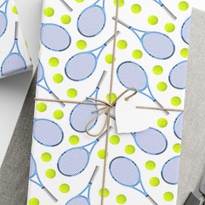 May include: Gift wrapped in white paper with a repeating pattern of blue tennis rackets and yellow tennis balls. The package is tied with twine and a blank white tag. A second wrapped gift is visible in the background.