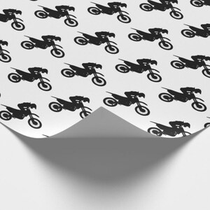 Op de afbeelding: Wit cadeaupapier met een herhaald patroon van zwarte motorfiets silhouetten. Het papier is gedeeltelijk gevouwen en onthult een witte onderkant en een schaduw.